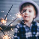 a small child holding a sparkler. The sparkler is clear in the foreground; the boy, who is wearing a grey hat and a blue plaid button-up shirt, is blurred in the background
